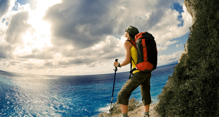 Woman hiking with trekking poles, standing on a seaside cliff admiring the view